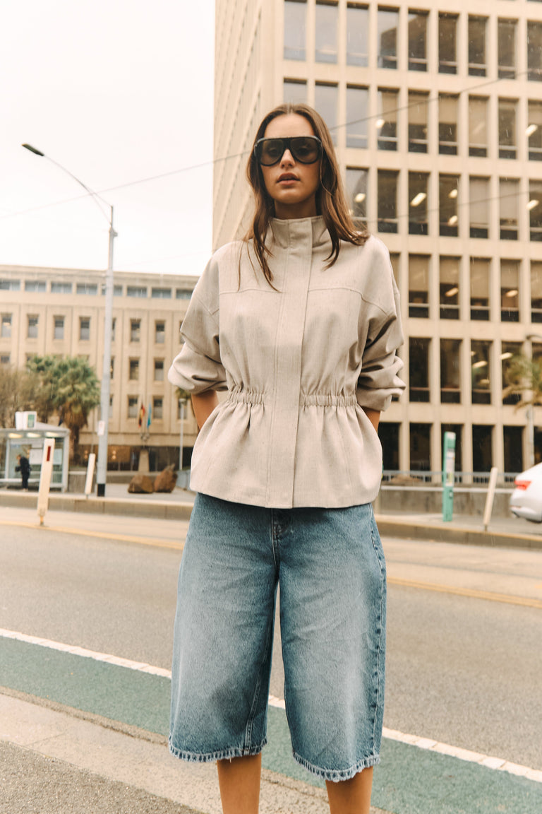 Woman wearing a beige gathered waist top and long light blue denim shorts with sunglasses outdoors