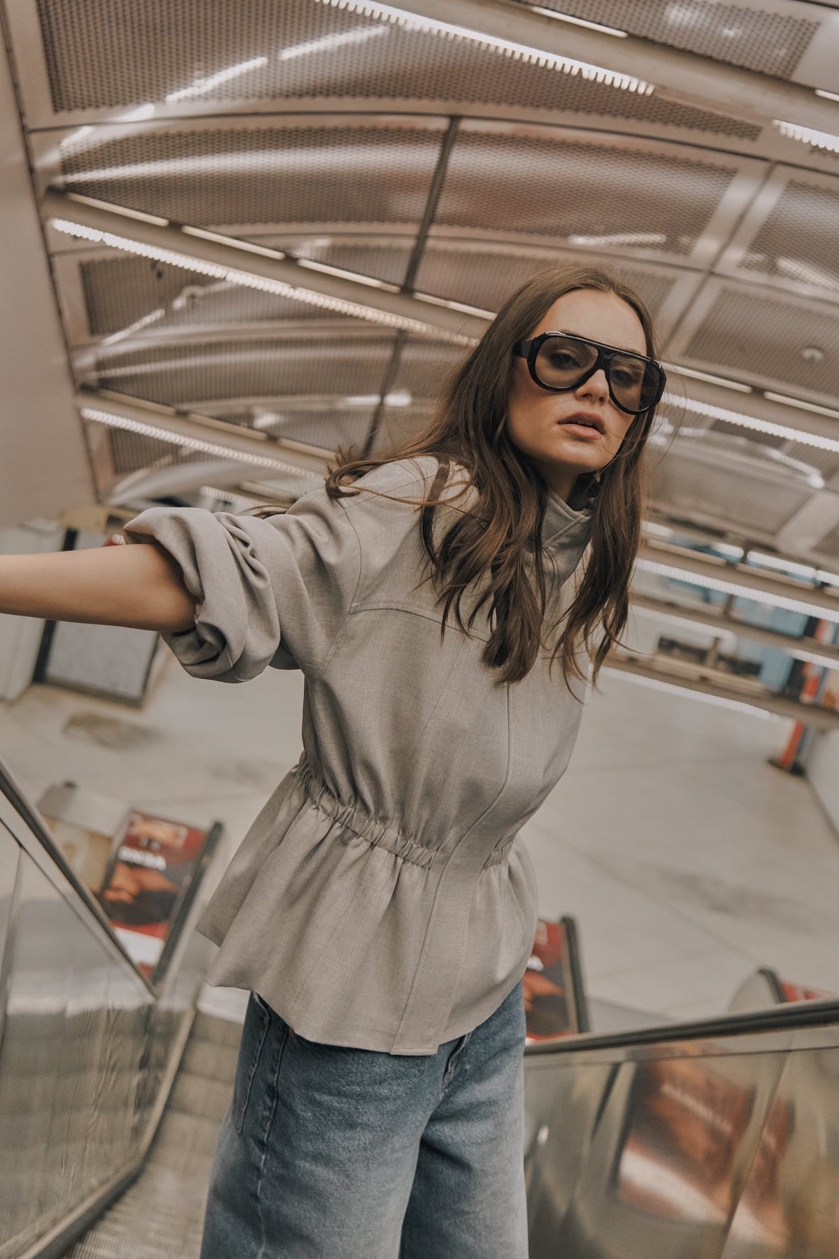 Woman wearing a grey cinched jacket with jeans and oversized sunglasses on an escalator