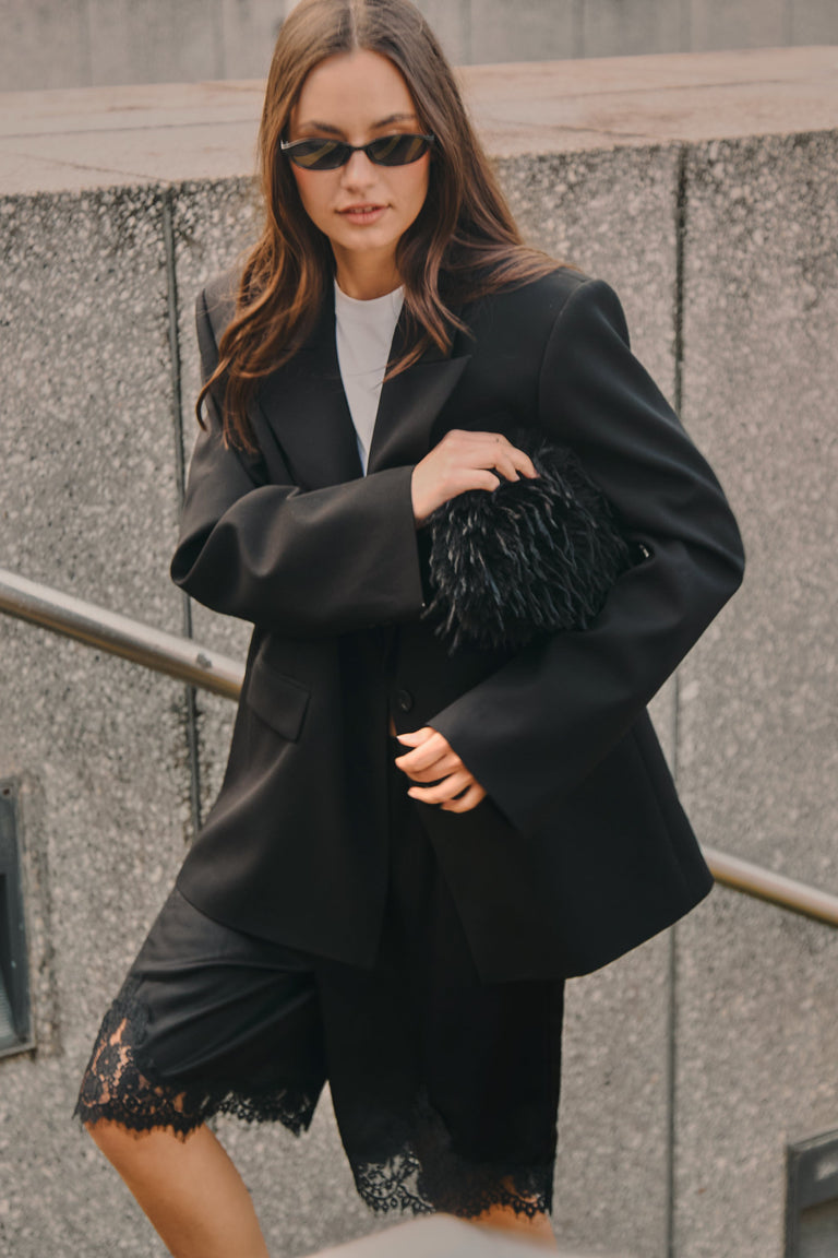 Woman wearing a black blazer with lace-trim satin Bermuda shorts, white tee, sunglasses and a textured clutch