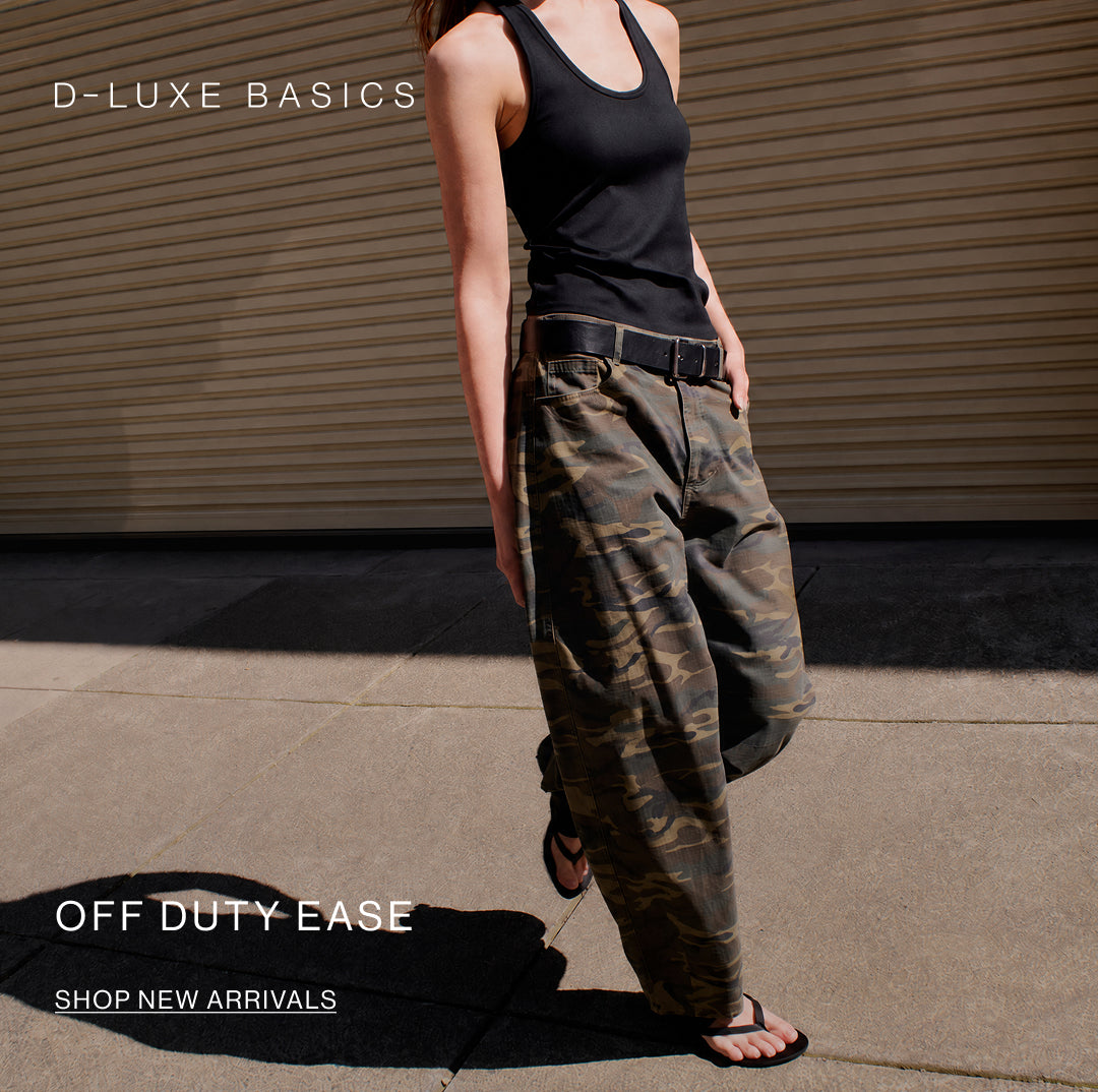 A model wearing a black tank top and wide-leg camouflage cargo pants with a black belt, walking outdoors in front of a corrugated metal wall. Text overlays read 'D-LUXE BASICS' and 'OFF DUTY EASE SHOP NEW ARRIVALS'.
