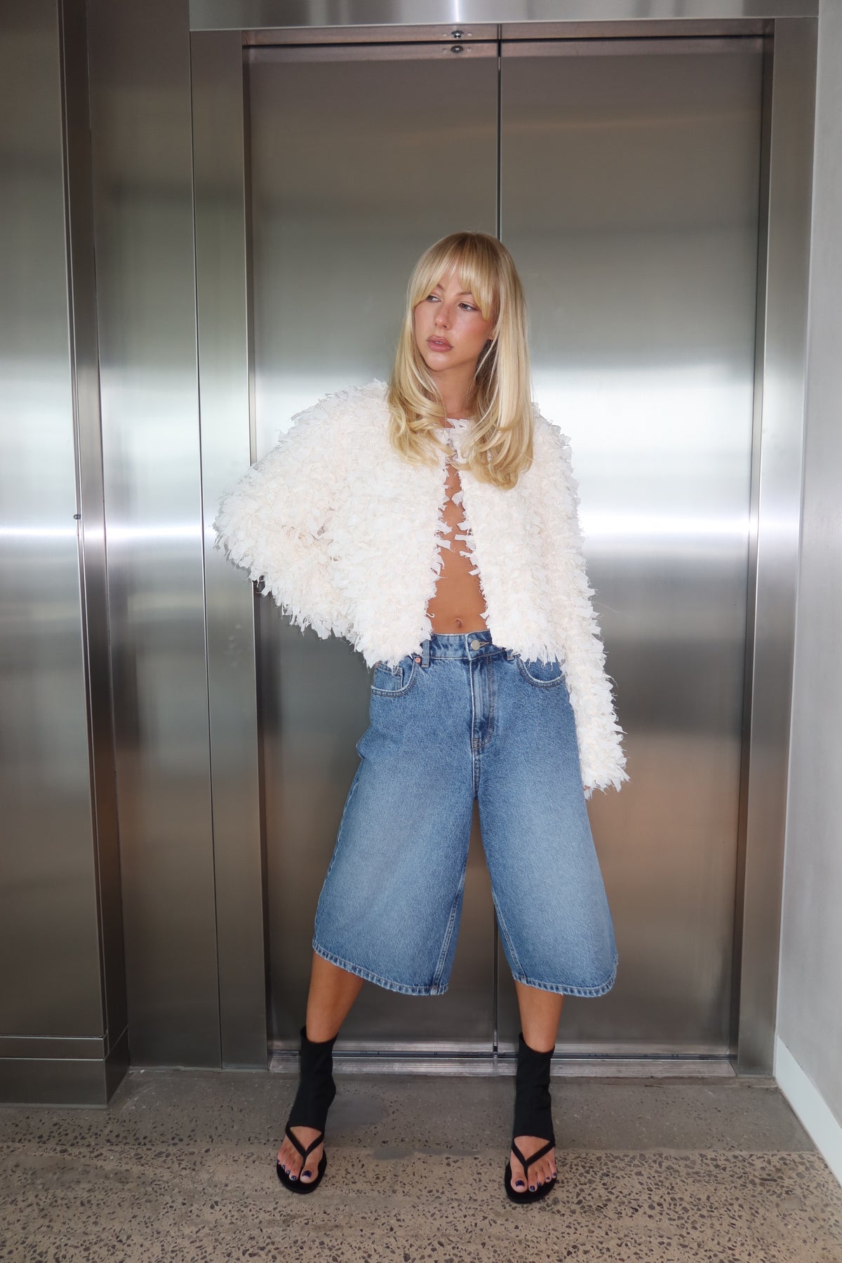 Woman wearing a white textured jacket with wide leg denim shorts and black sandals