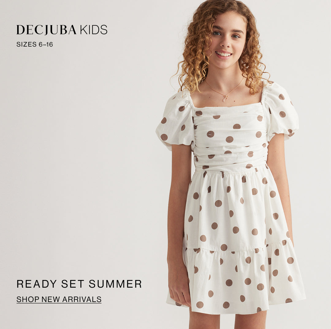 A young girl wearing a Decjuba Kids white dress with brown polka dots, featuring puff sleeves and a tiered skirt, set against a plain light background with promotional text.