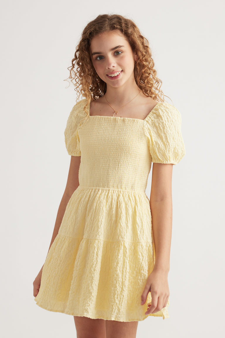 Girl wearing a pale yellow textured mini dress with a shirred bodice, square neckline and puff sleeves