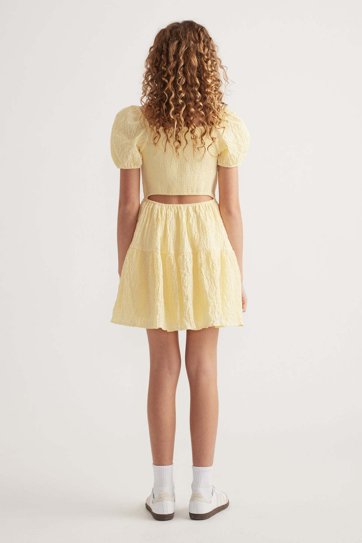 Girl seen from behind in a yellow textured mini dress with puff sleeves, back cutout and sneakers
