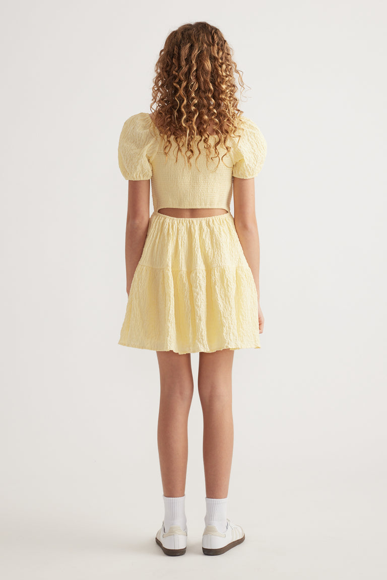 Girl seen from behind in a yellow textured mini dress with puff sleeves, back cutout and sneakers