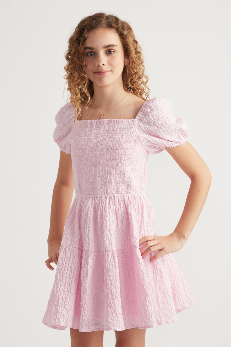 Girl wearing a pink textured mini dress with a shirred bodice square neckline and puff sleeves