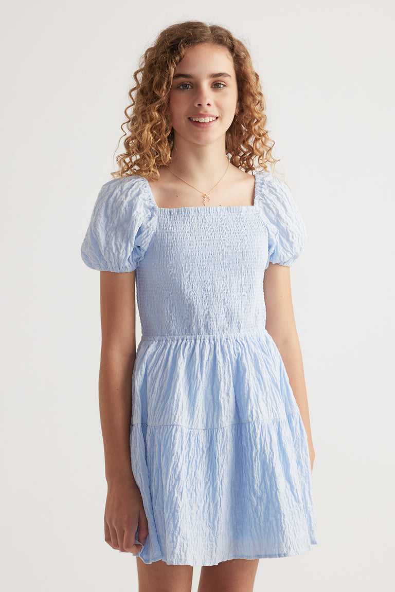 Girl wearing a light blue textured mini dress with a shirred bodice, square neckline and puff sleeves