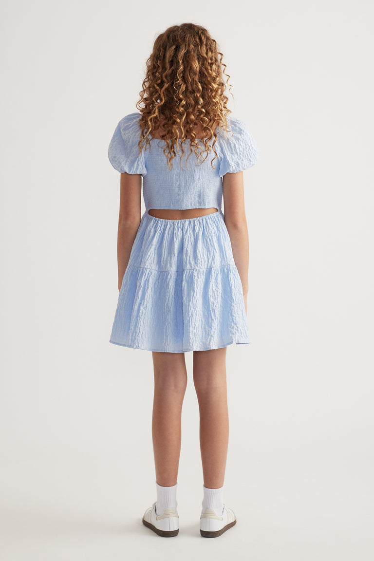 Girl seen from behind wearing a light blue textured puff sleeve dress with a back cutout and white sneakers