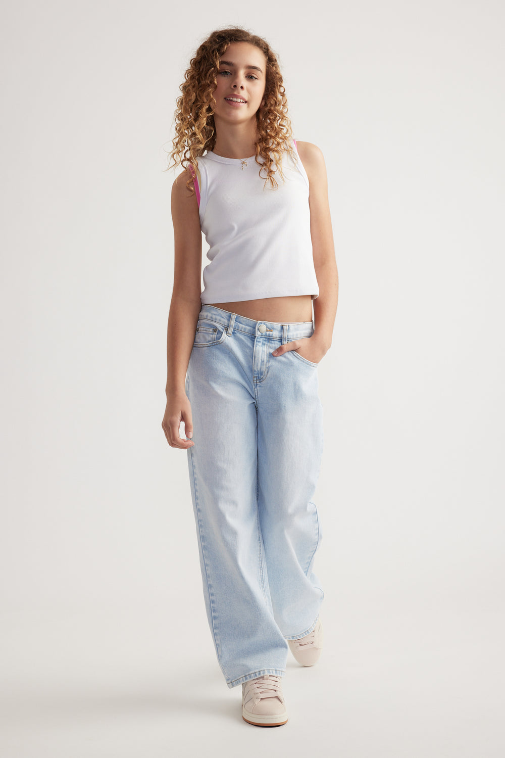 Teen girl wearing light wash baggy mid-rise jeans with a sleeveless white crop top and sneakers against a plain background