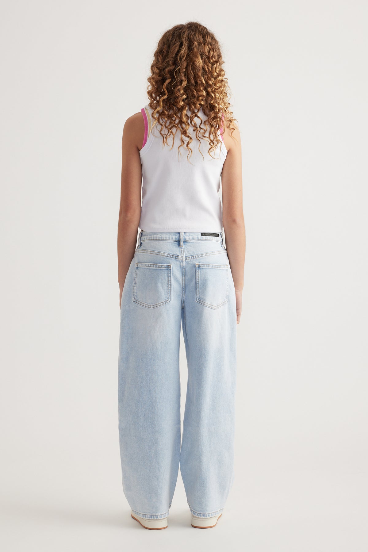 Rear view of girl wearing light wash baggy fit mid-rise jeans with a white sleeveless top against plain background
