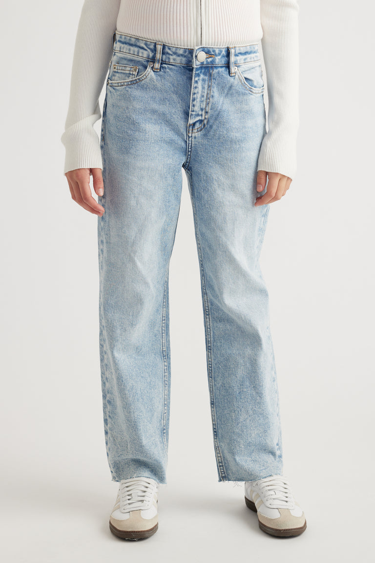 Front view of girl wearing light wash straight leg jeans with raw hems paired with white sneakers and cream jumper