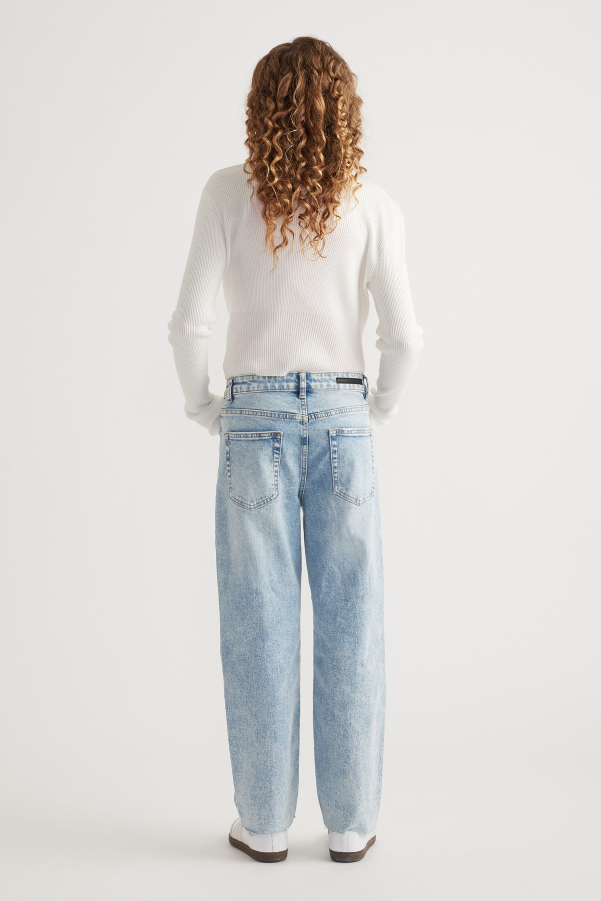 Rear view of girl wearing light wash straight leg jeans with raw hem paired with white long sleeve top