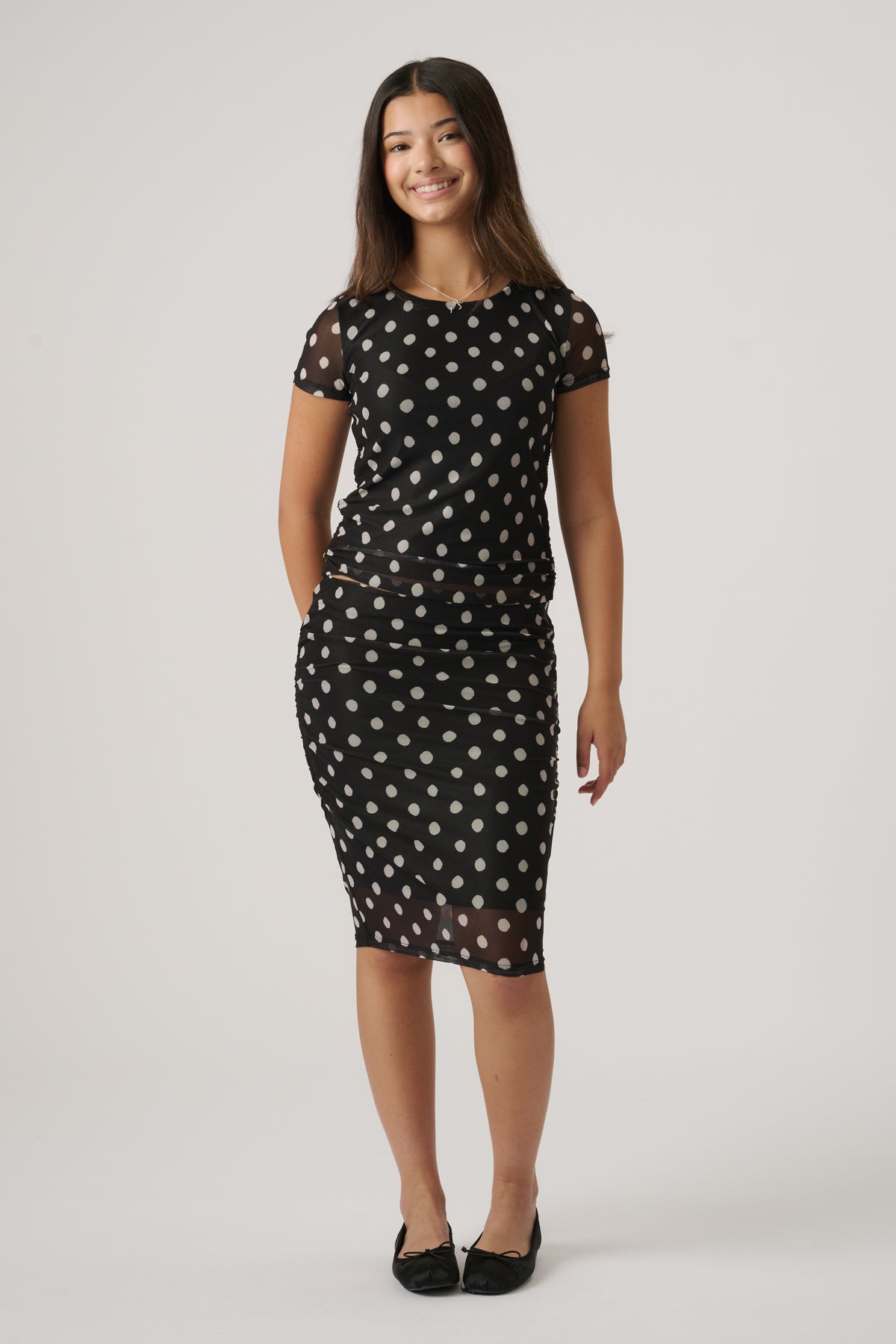 Teen girl wearing sheer black top and matching midi skirt with white polka dots and ruching details