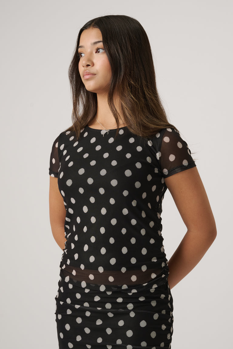 Young girl wearing a black mesh top with white polka dots and short sleeves, styled with matching black skirt