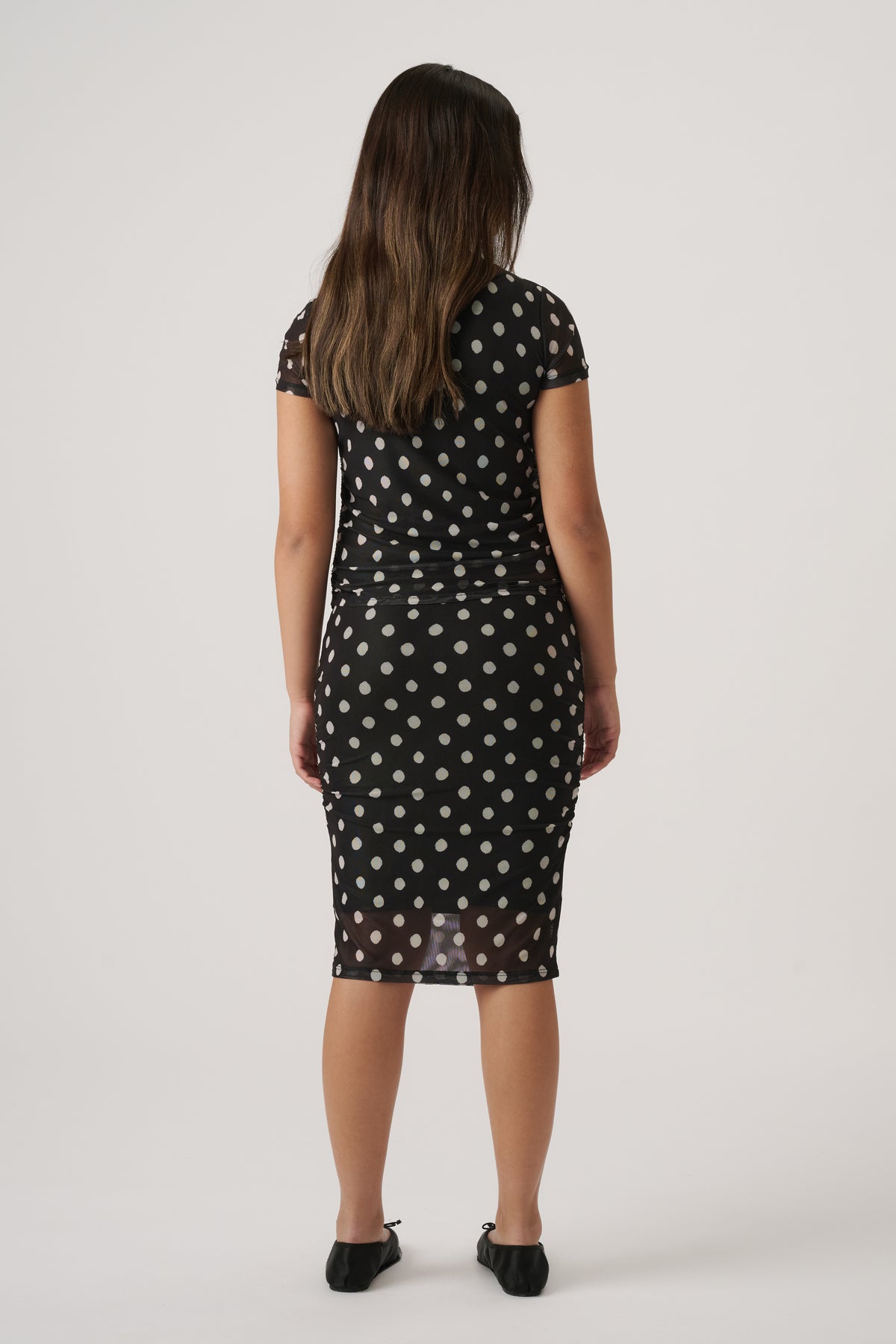 Girl wearing a black mesh dress with white polka dots and short sleeves, paired with black ballet flats, facing away