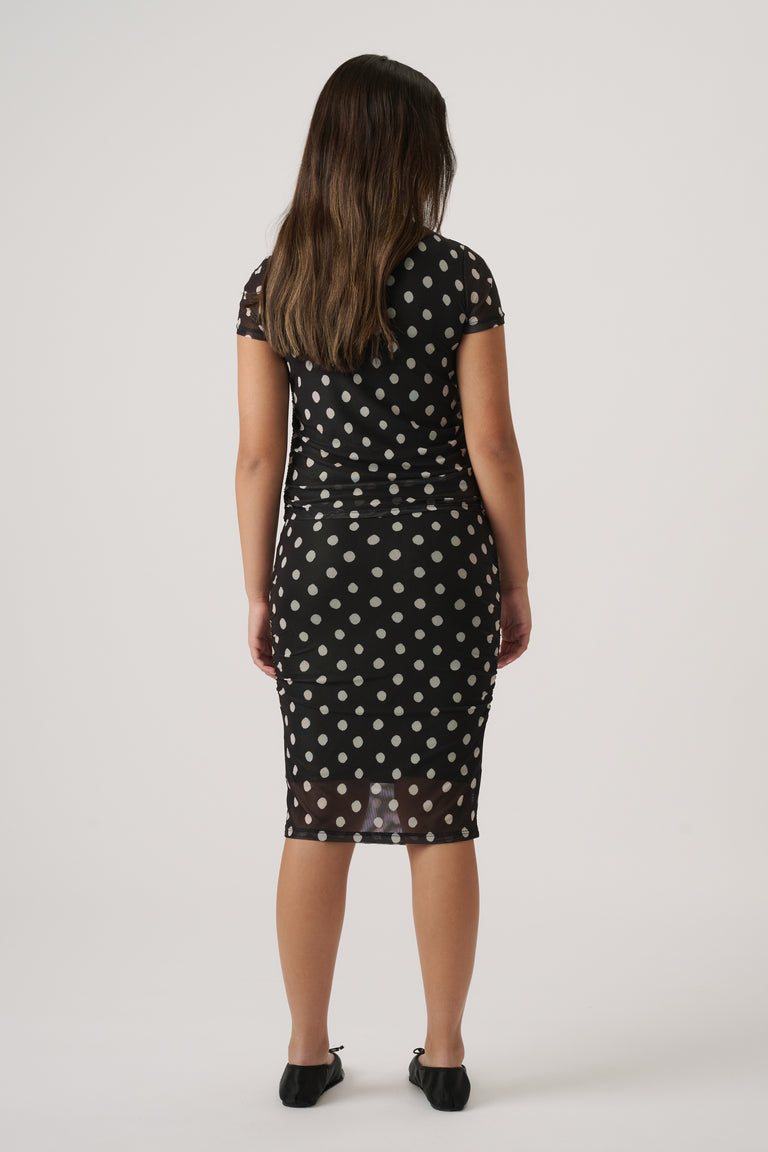 Girl wearing a black mesh dress with white polka dots and short sleeves, paired with black ballet flats, facing away