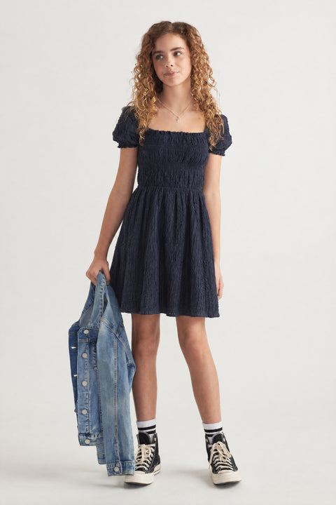 Girl wearing a navy textured mini dress with puff sleeves, denim jacket and high top sneakers