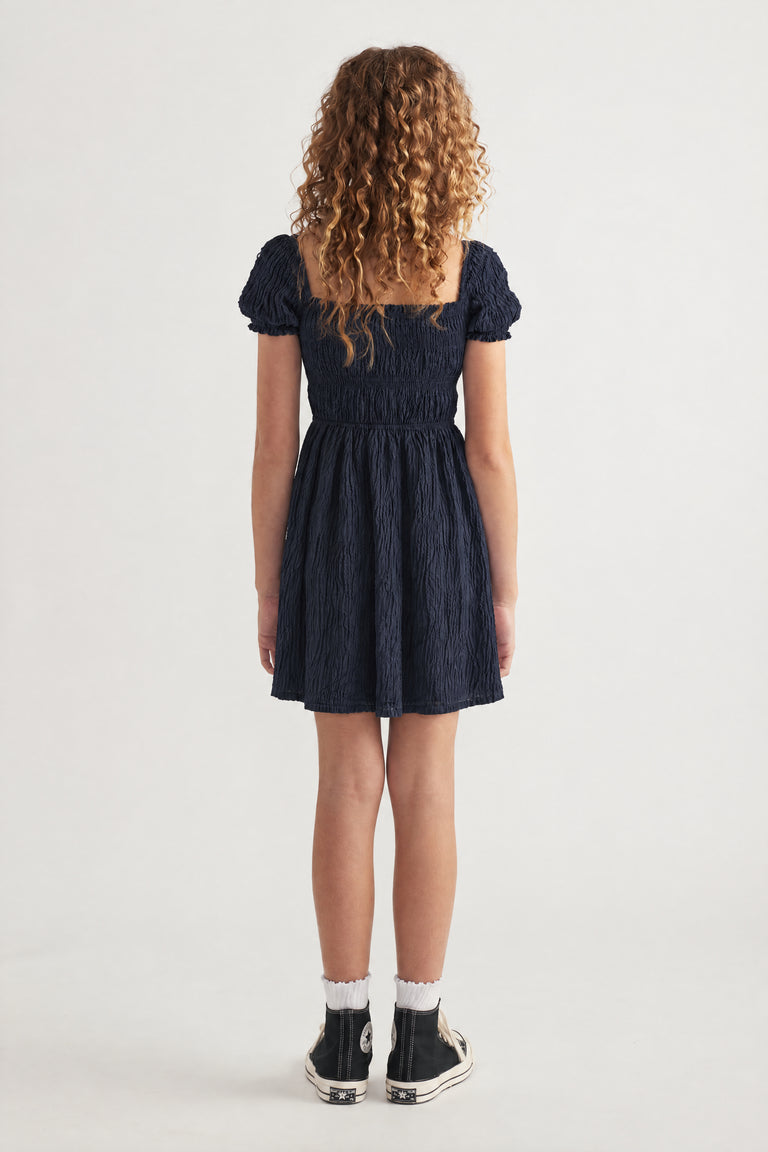 Girl wearing a navy textured mini dress with puff sleeves and tiered skirt, viewed from behind