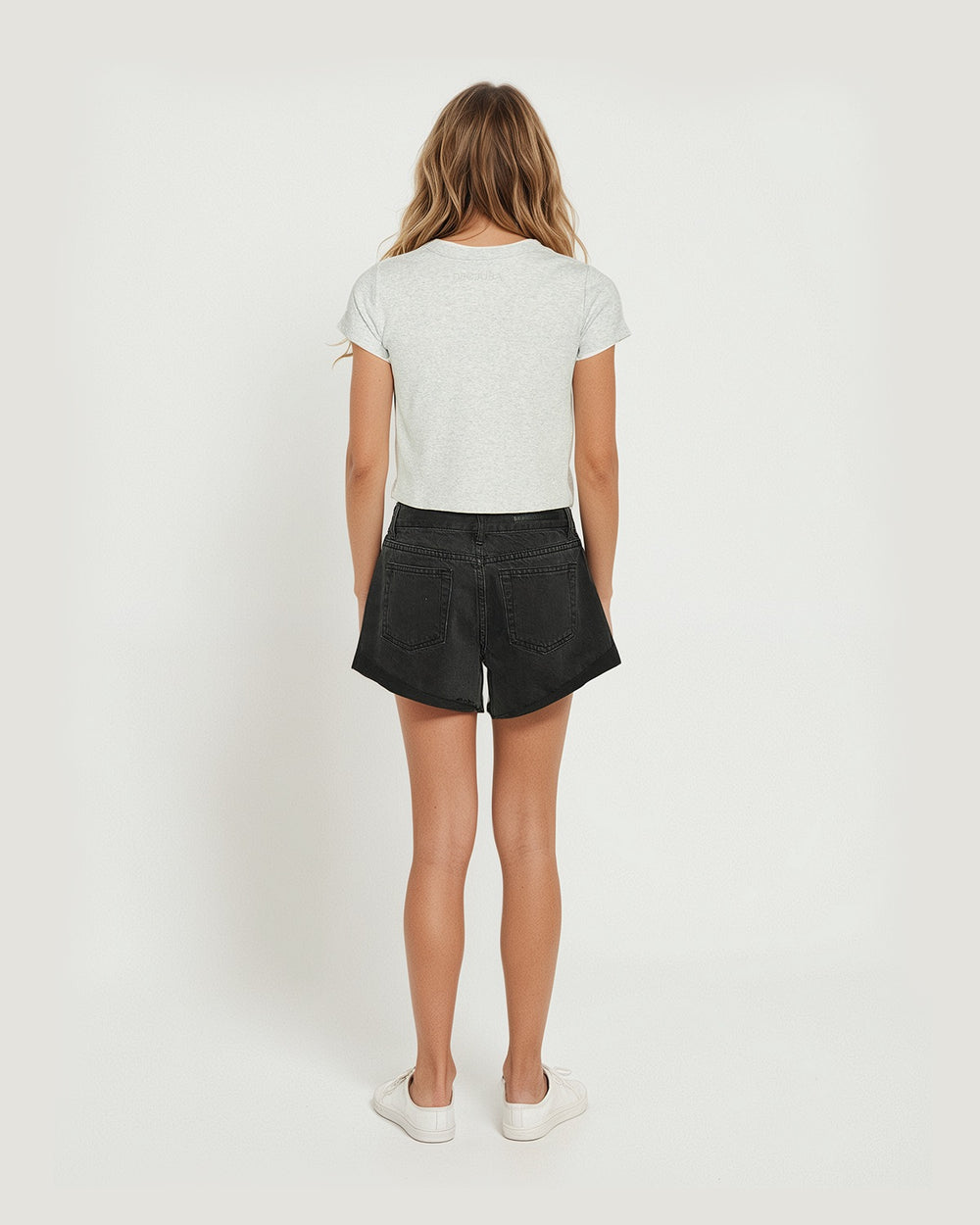 Young girl seen from behind wearing washed black denim shorts paired with a simple light grey tee and white sneakers.