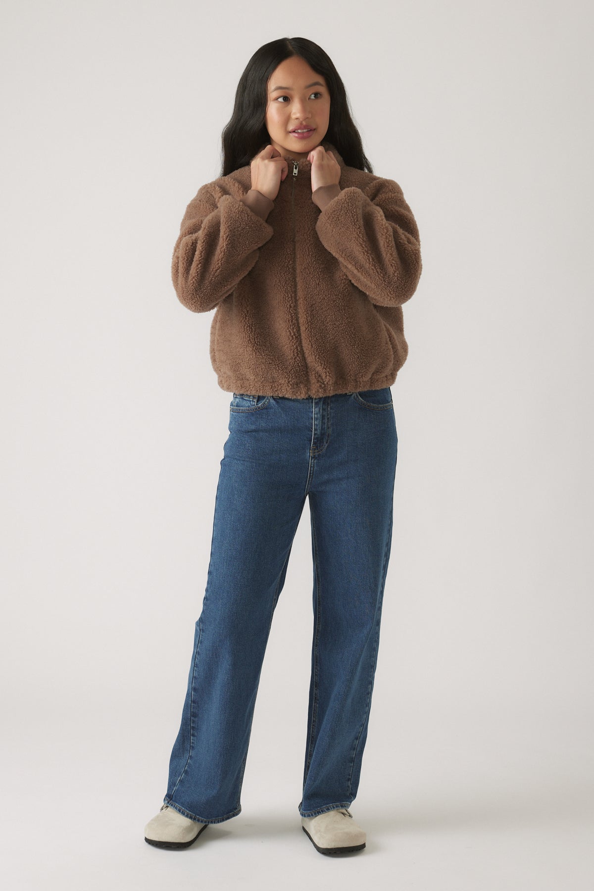 Teen girl wearing relaxed fit blue jeans paired with a cozy brown zip-up teddy jacket and casual slip-on shoes