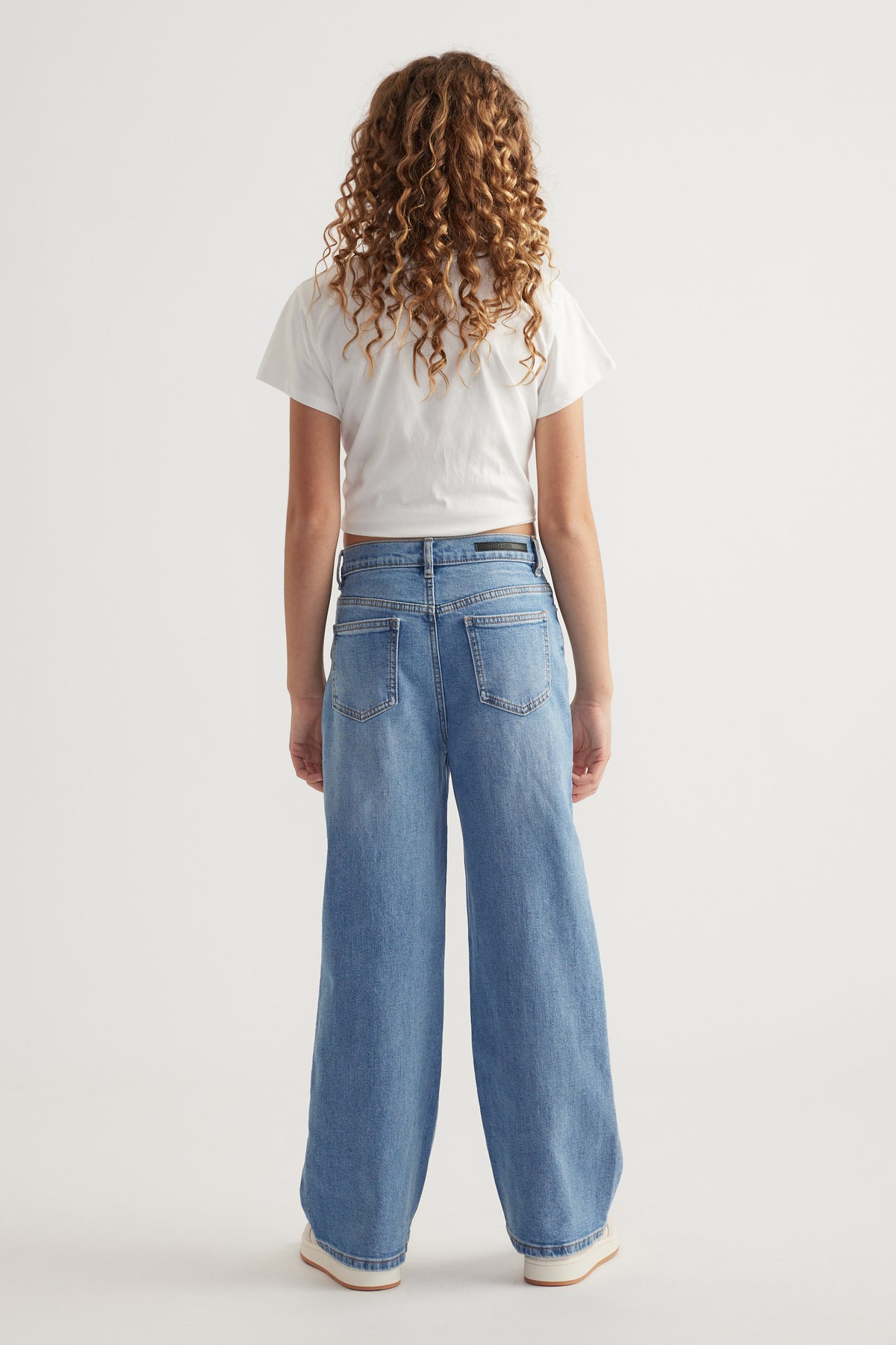Rear view of girl wearing relaxed fit, mid-rise light blue jeans with functional back pockets and a white cropped tee