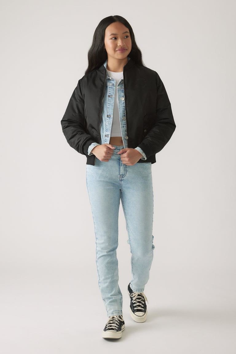 Teen girl wearing light wash skinny jeans layered with a denim jacket and black cropped bomber jacket with sneakers