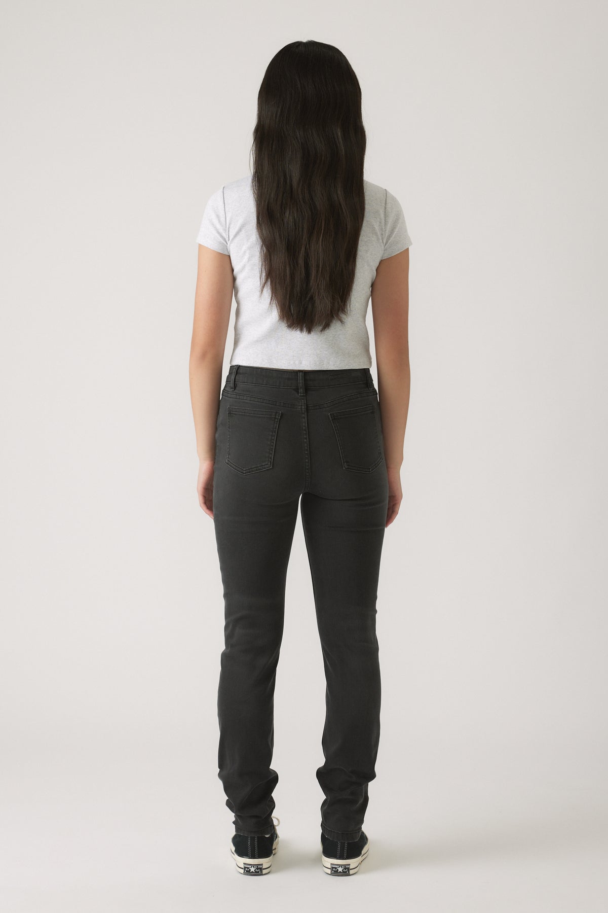 Girl wearing dark skinny jeans paired with a fitted light grey t-shirt and black sneakers, shown from the back