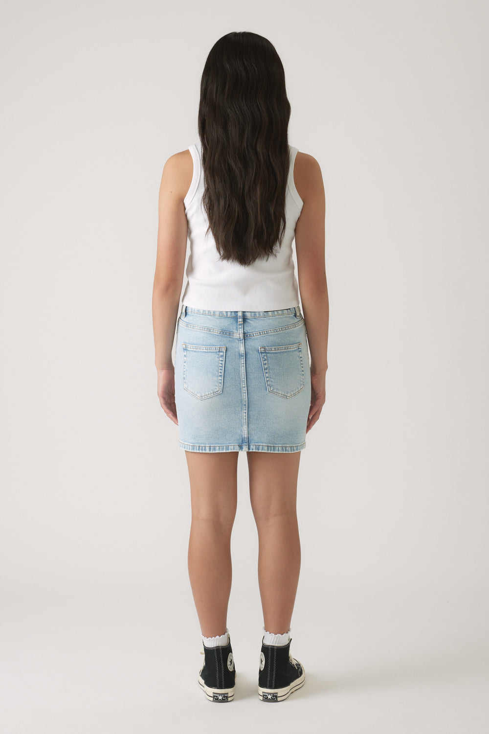 Girl wearing a light wash denim skirt paired with a sleeveless white top and black high-top sneakers, back view.