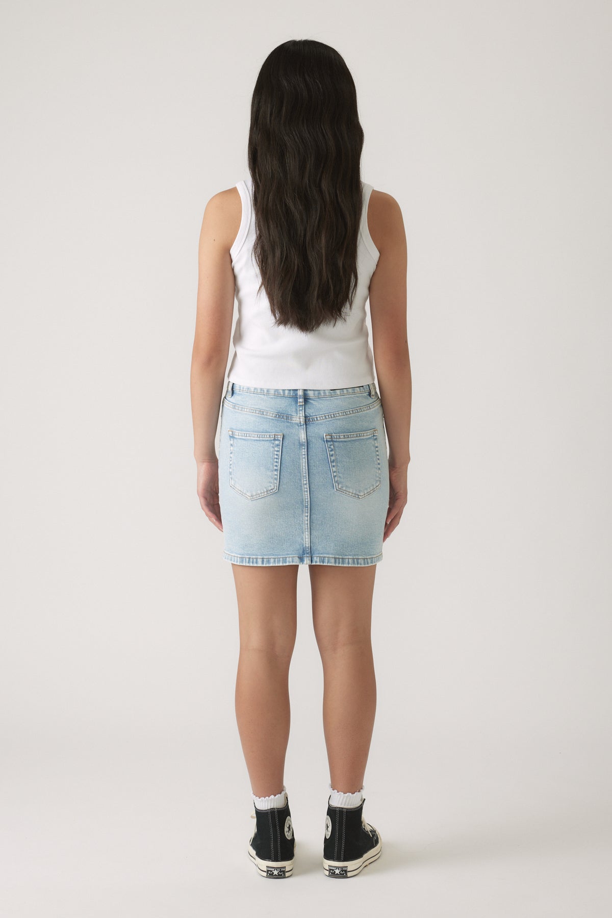 Girl wearing a light wash denim skirt paired with a sleeveless white top and black high-top sneakers, back view.