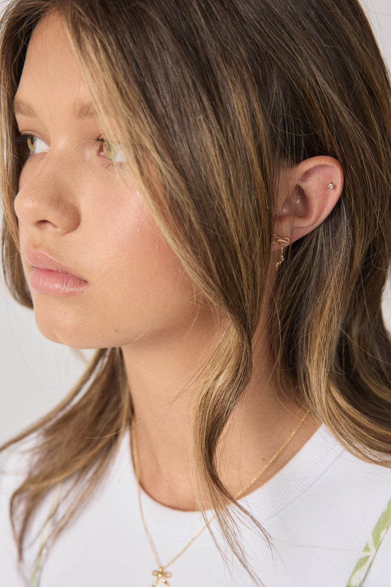 Close-up of a girl wearing delicate gold bow stud earrings with layered gold jewelry and soft wavy hair
