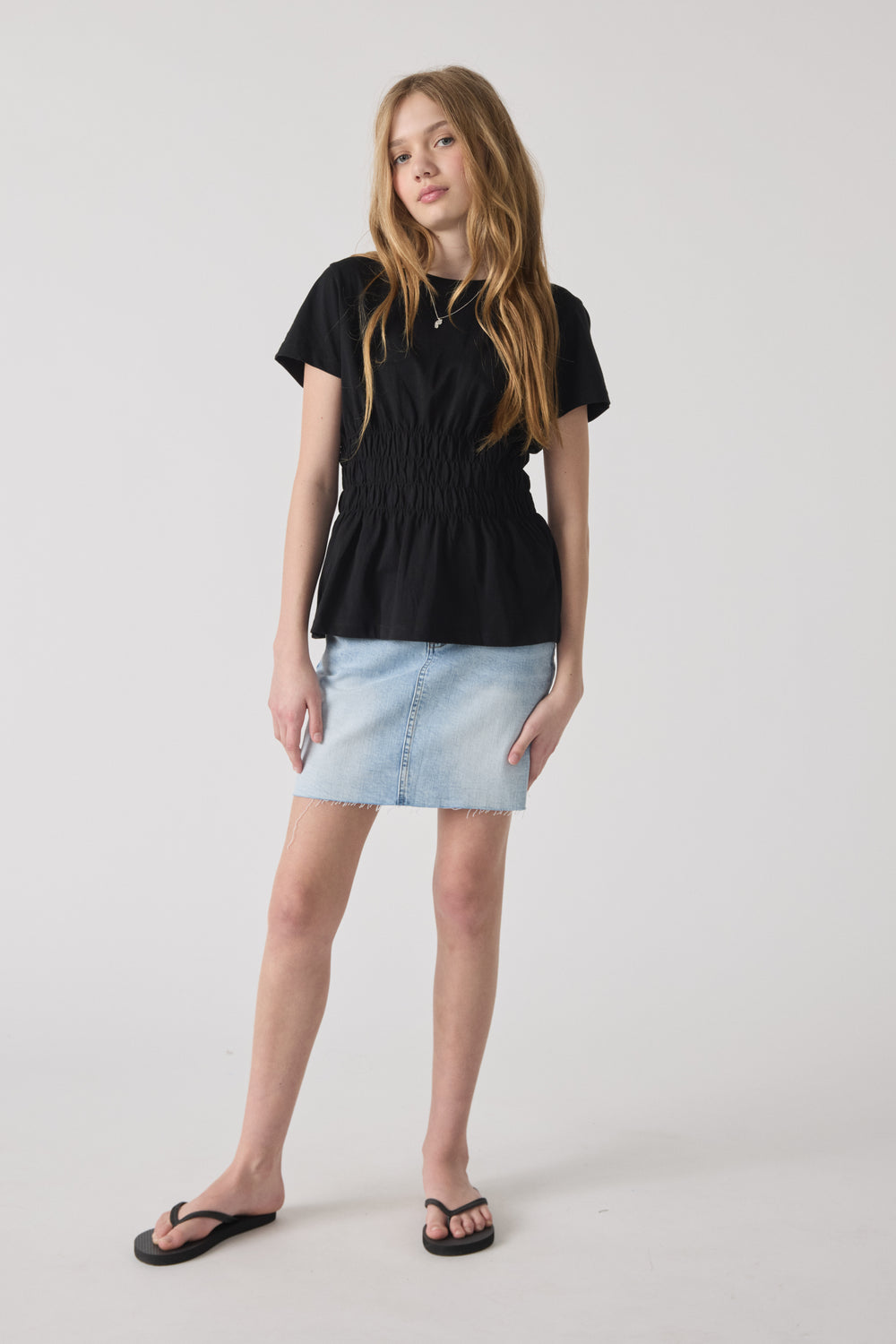 Girl wearing a fitted black smocked tee paired with a light blue denim skirt and black flip-flops against a neutral backdrop