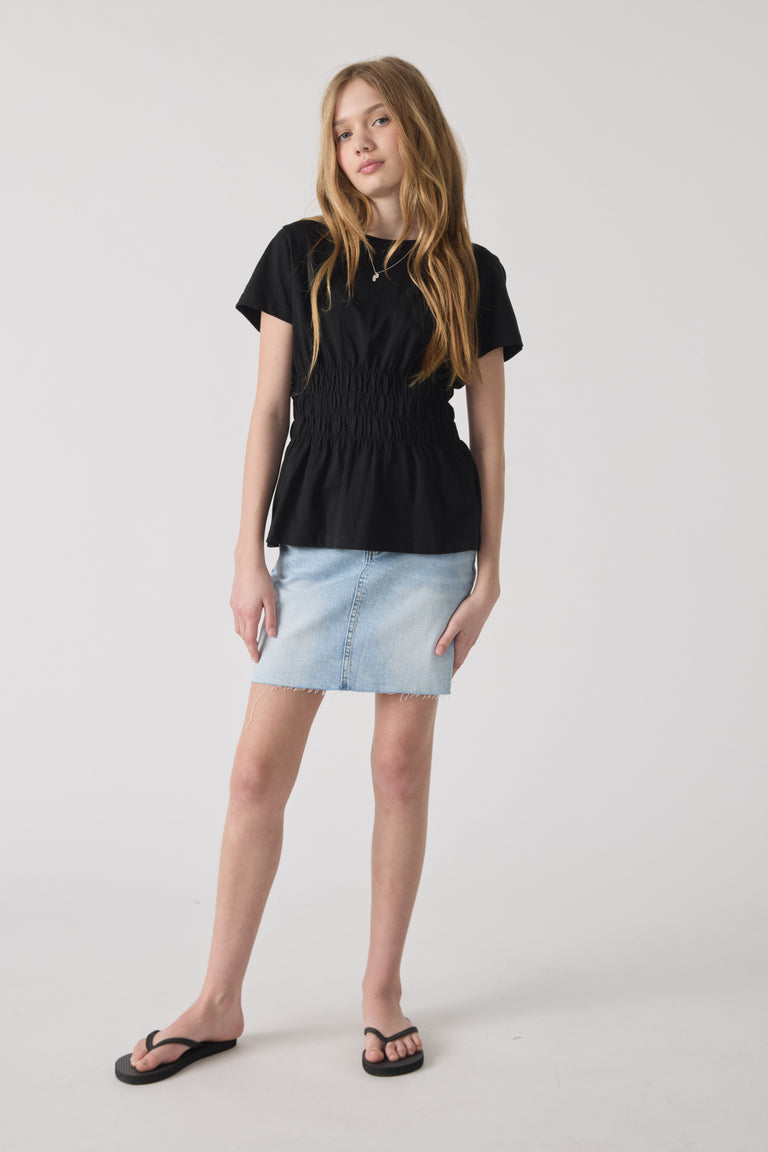 Girl wearing a fitted black smocked tee paired with a light blue denim skirt and black flip-flops against a neutral backdrop