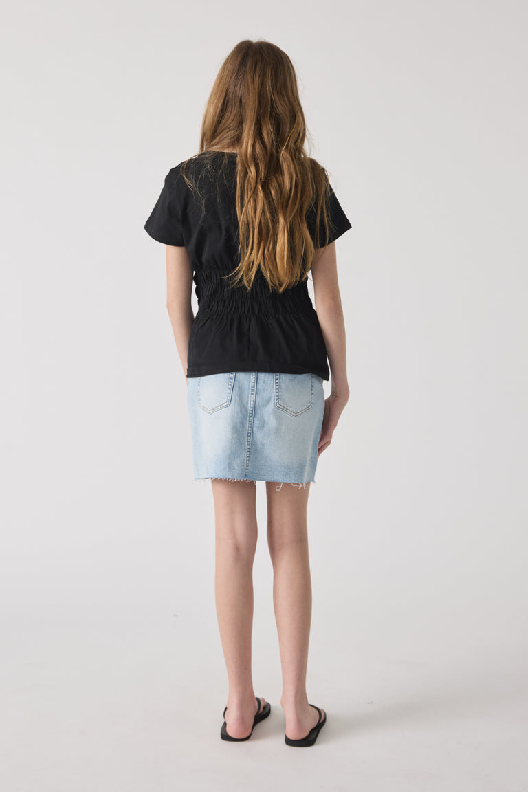 Rear view of girl wearing a black smocked tee paired with a light wash denim skirt and black sandals