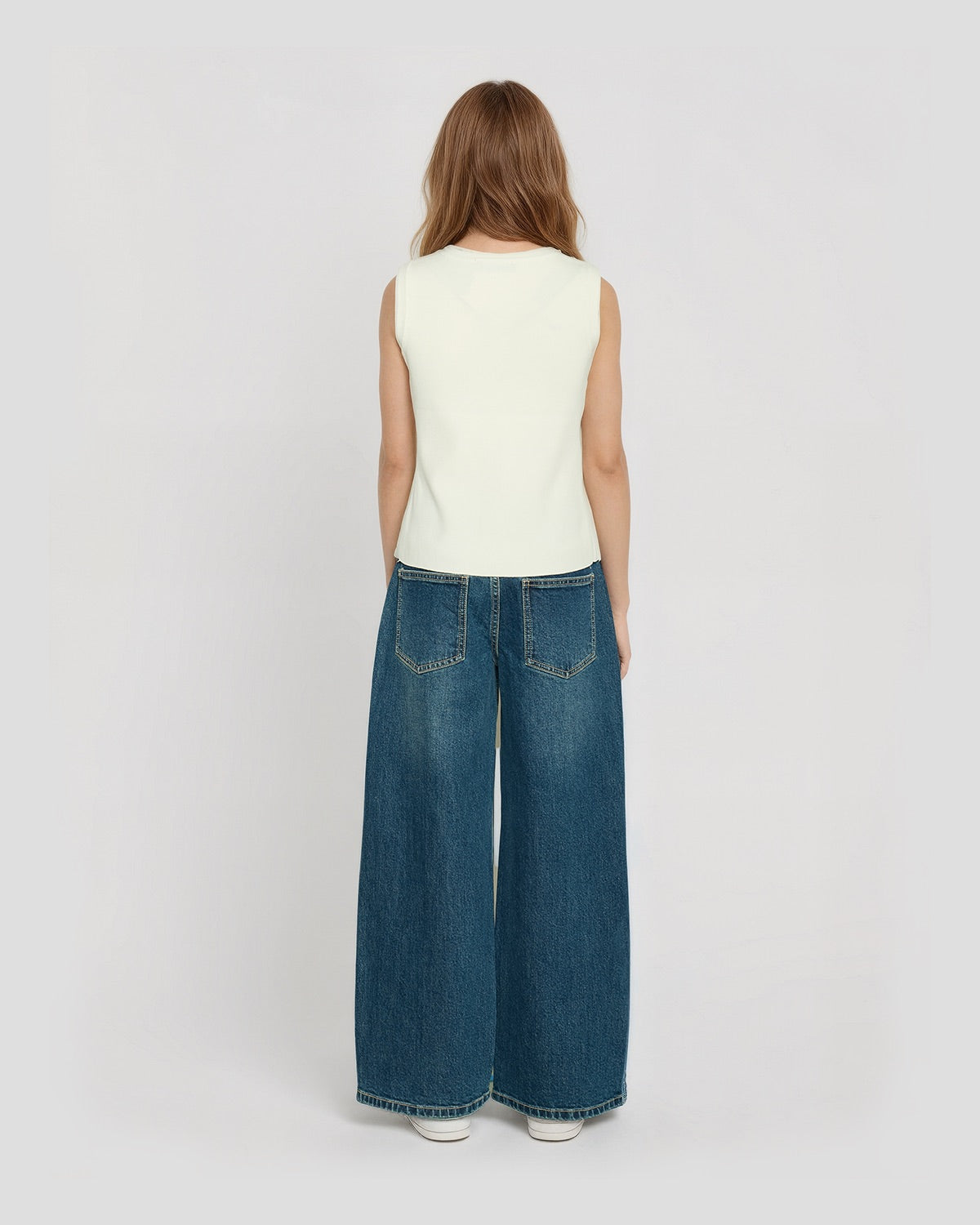 Back view of girl wearing a cream sleeveless knit vest paired with wide-leg blue denim jeans and white sneakers.