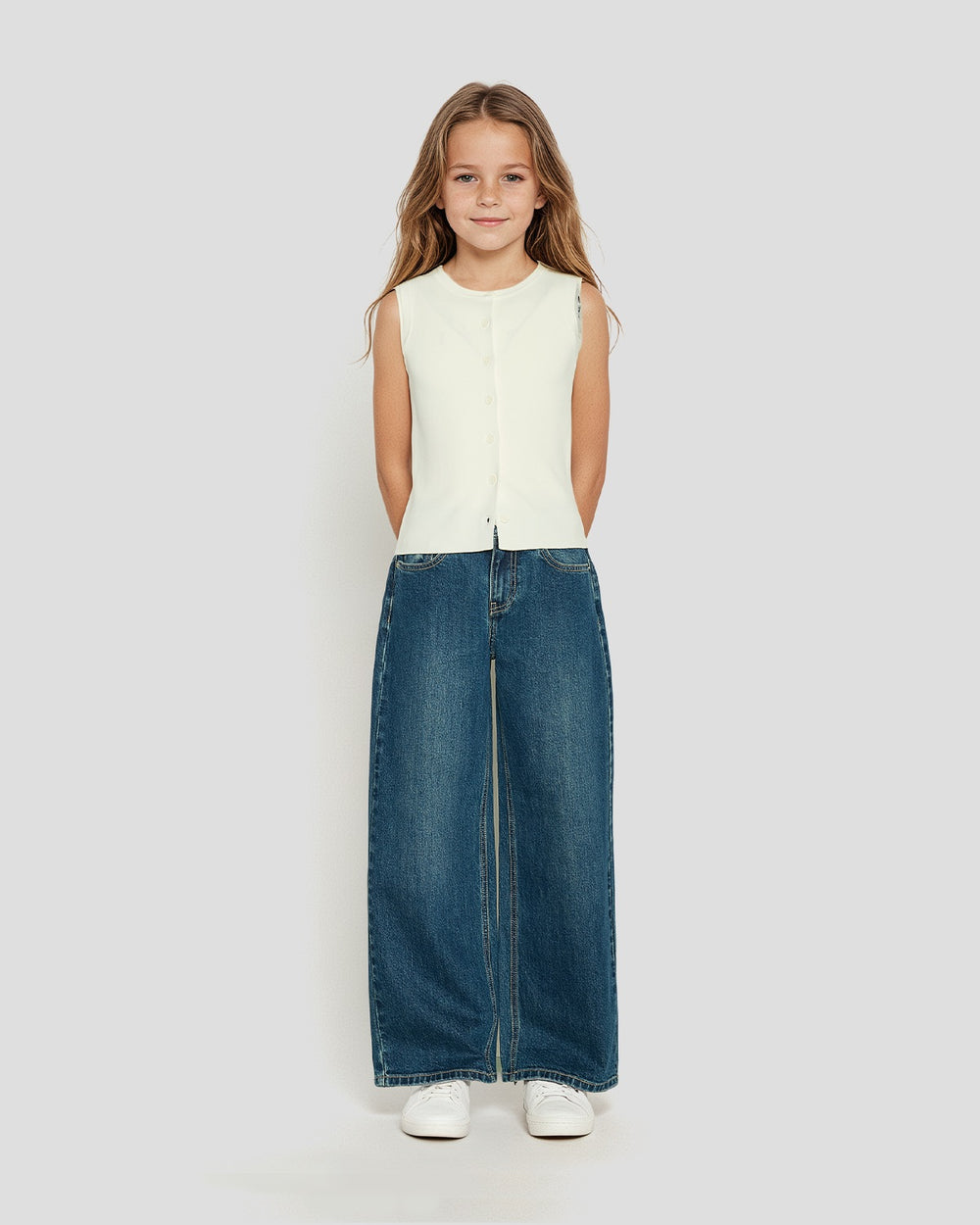 Young girl wearing a sleeveless cream knit vest paired with wide-leg blue denim jeans and white sneakers
