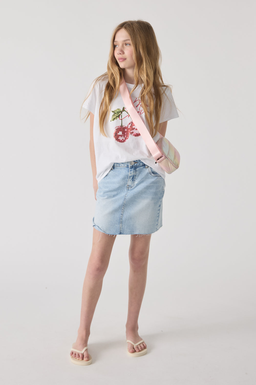 Girl wearing a white cherry graphic tee, light denim skirt, flip-flops, and a pastel padded crossbody bag with pink strap.