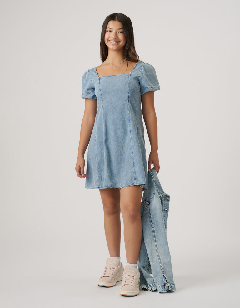 Girl wearing a light blue denim mini dress with puff sleeves, paired with sneakers and holding a matching denim jacket.