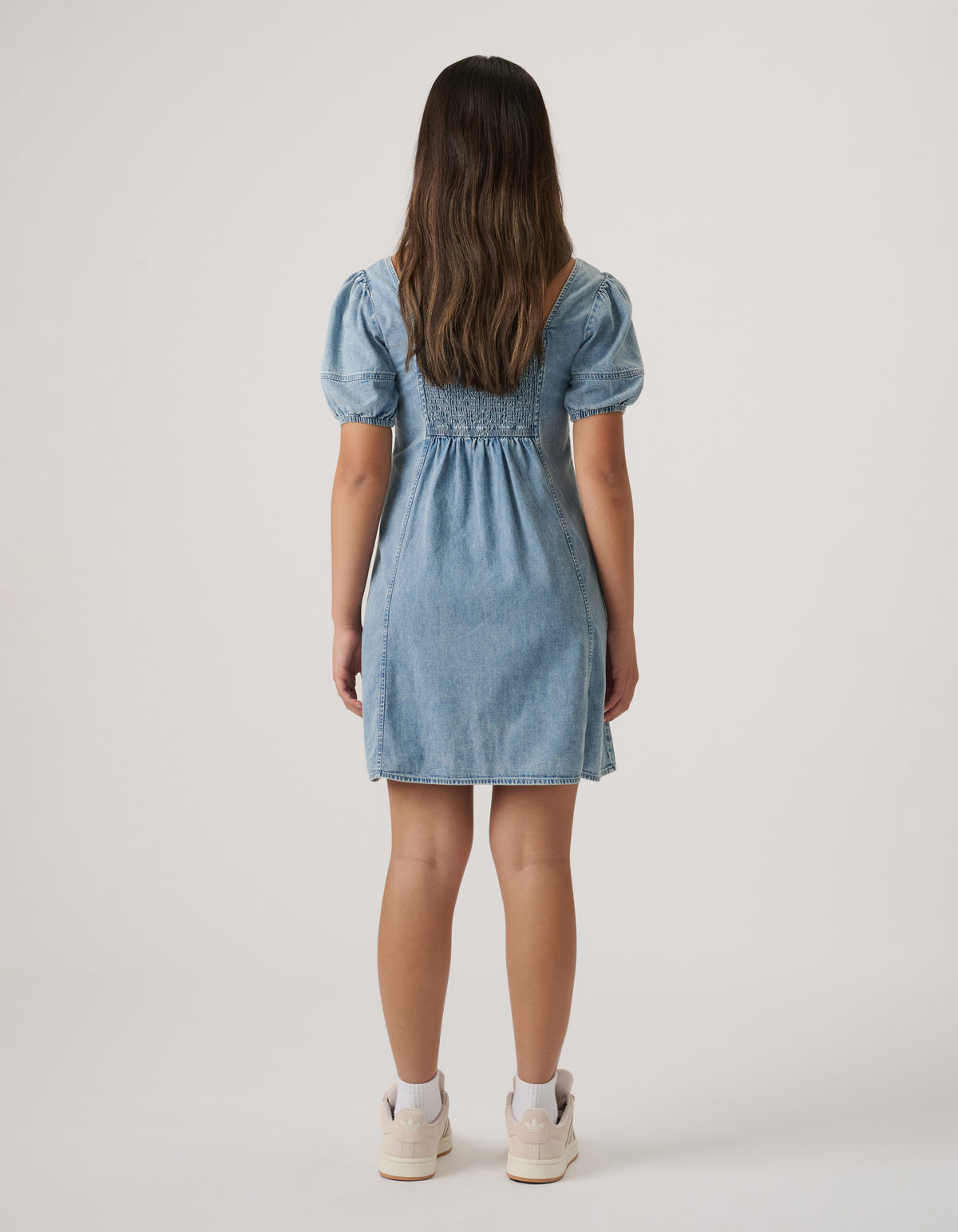 Girl wearing a light wash denim mini dress with puff sleeves and smocked back detail, paired with white sneakers and socks