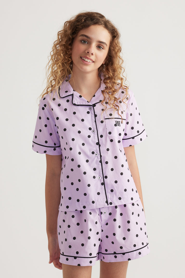 Girl wearing a lilac polka dot sleep set with a collared short sleeve shirt and matching shorts