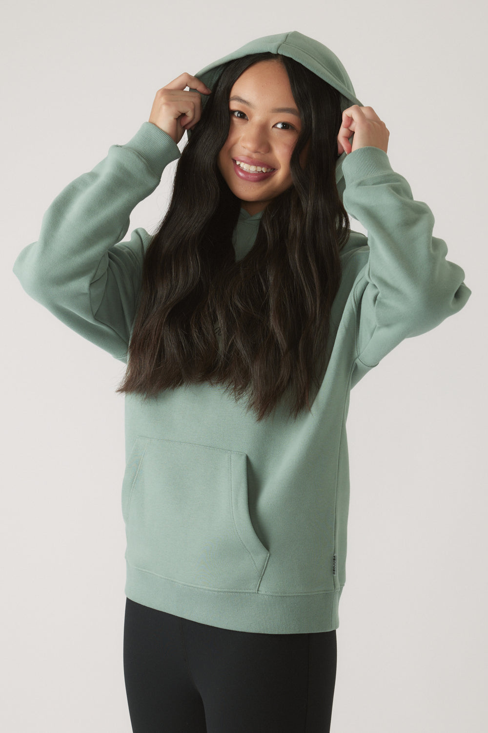 Smiling girl adjusting hood on moss green oversized hoodie paired with black leggings against a plain backdrop