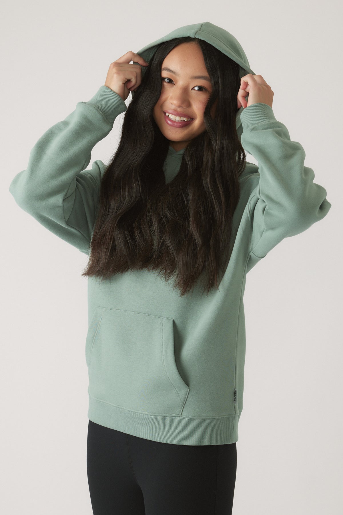Smiling girl adjusting hood on moss green oversized hoodie paired with black leggings against a plain backdrop
