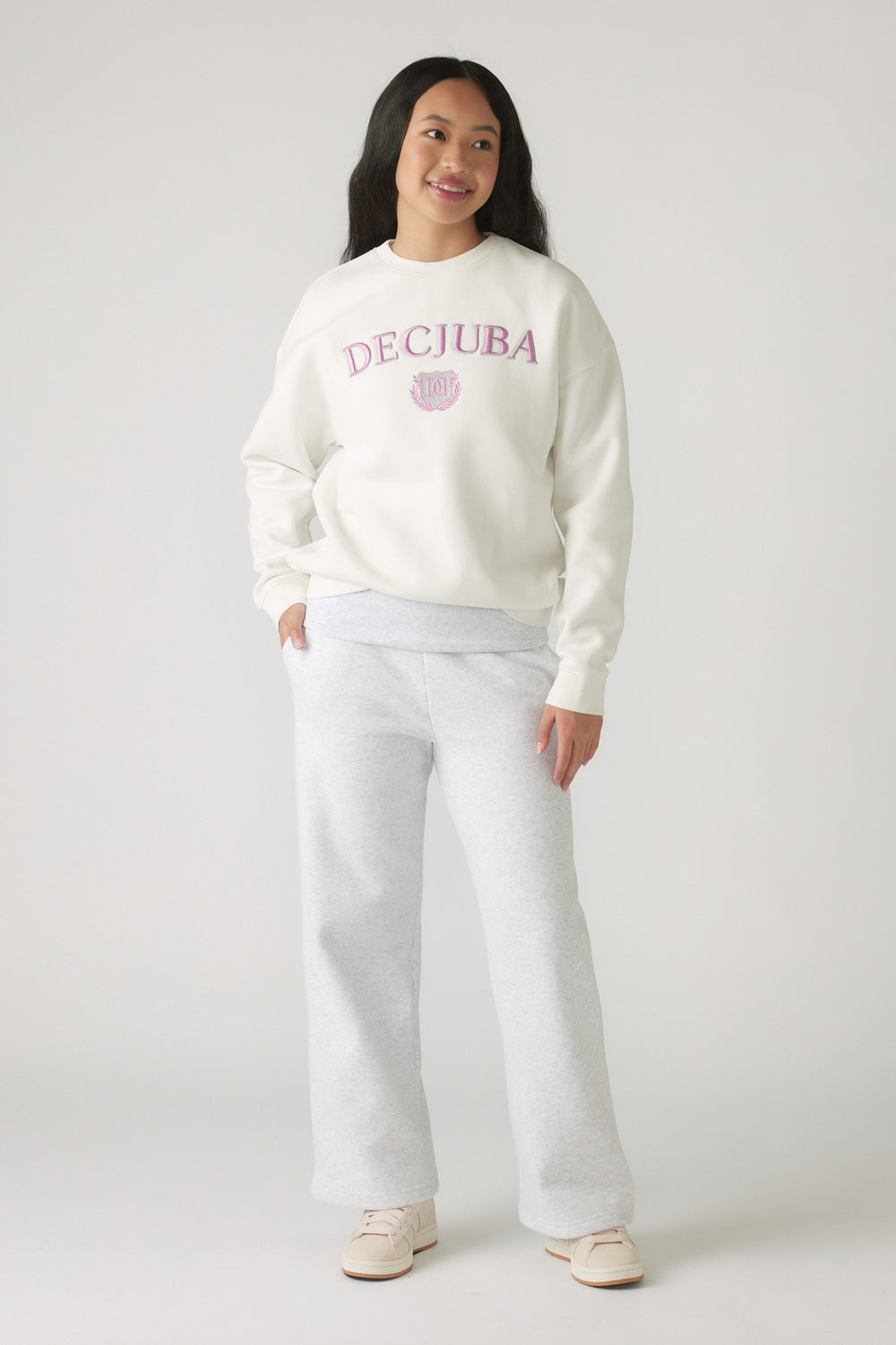 Teen girl smiling wearing a relaxed off-white sweatshirt with pink lettering paired with light grey wide-leg pants and cream sneakers