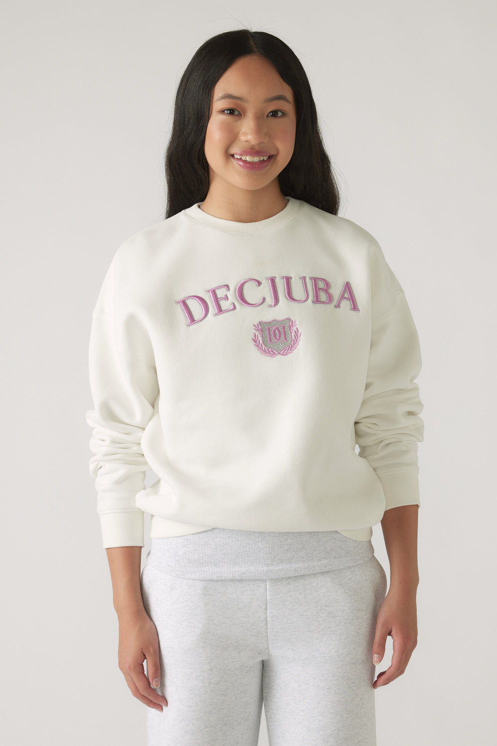 Teen girl smiling, wearing an oversized cream sweatshirt with pink lettering paired with light grey pants.
