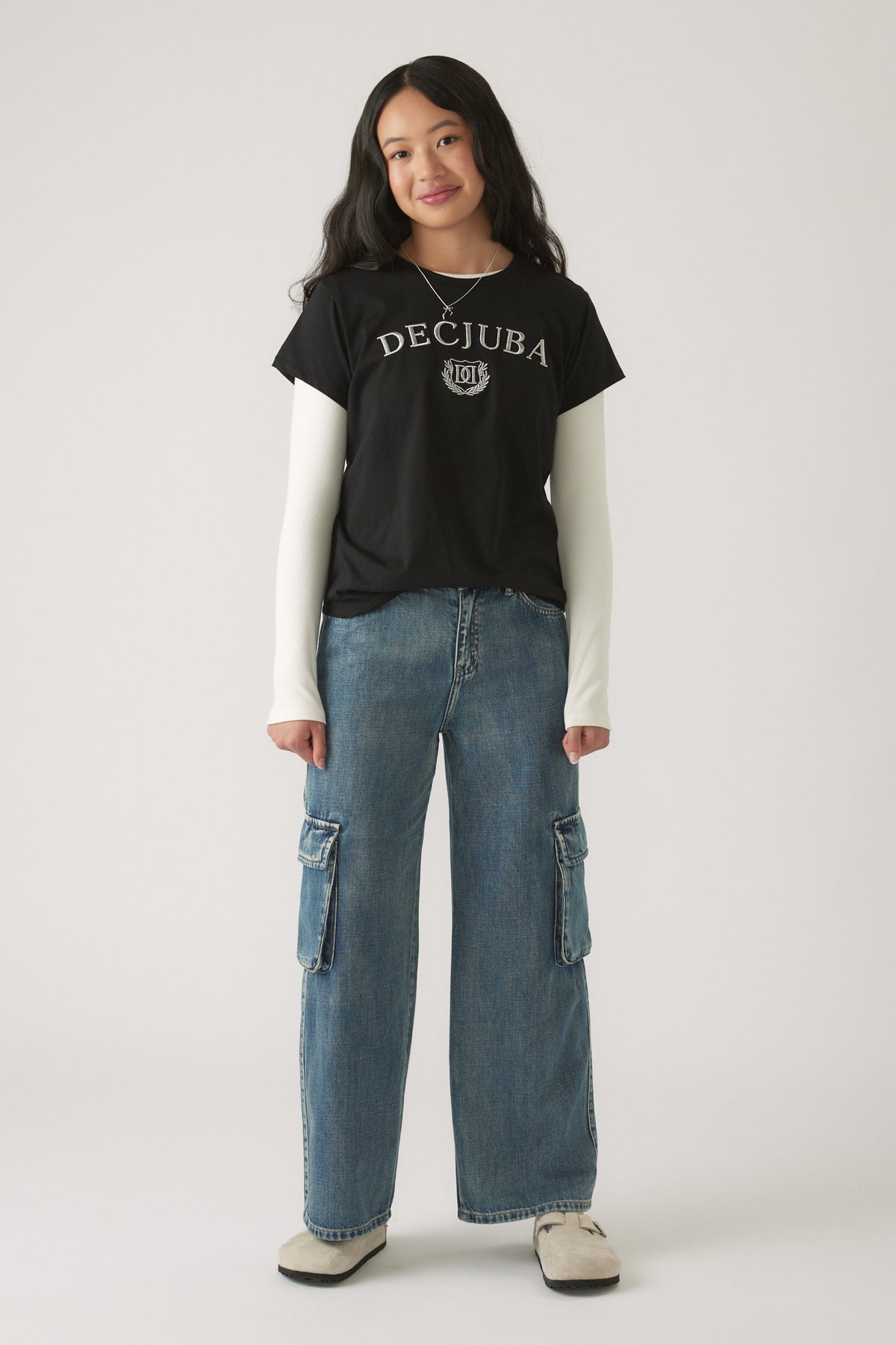 Girl wearing a Girls Decjuba Heritage Tee in black layered over white sleeves, styled with relaxed cargo denim jeans and sneakers