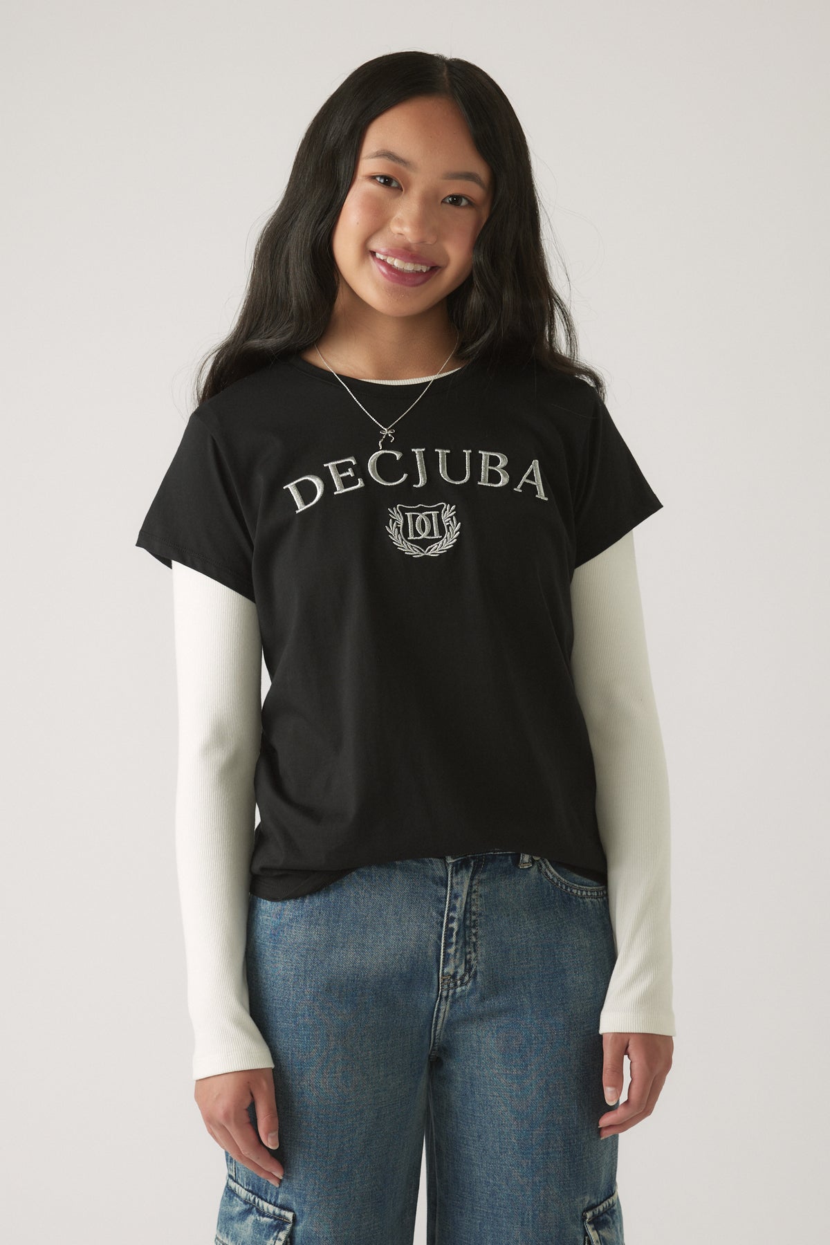 Girl smiling wearing a Girls Decjuba Heritage Tee in black layered over white long sleeves with relaxed denim jeans