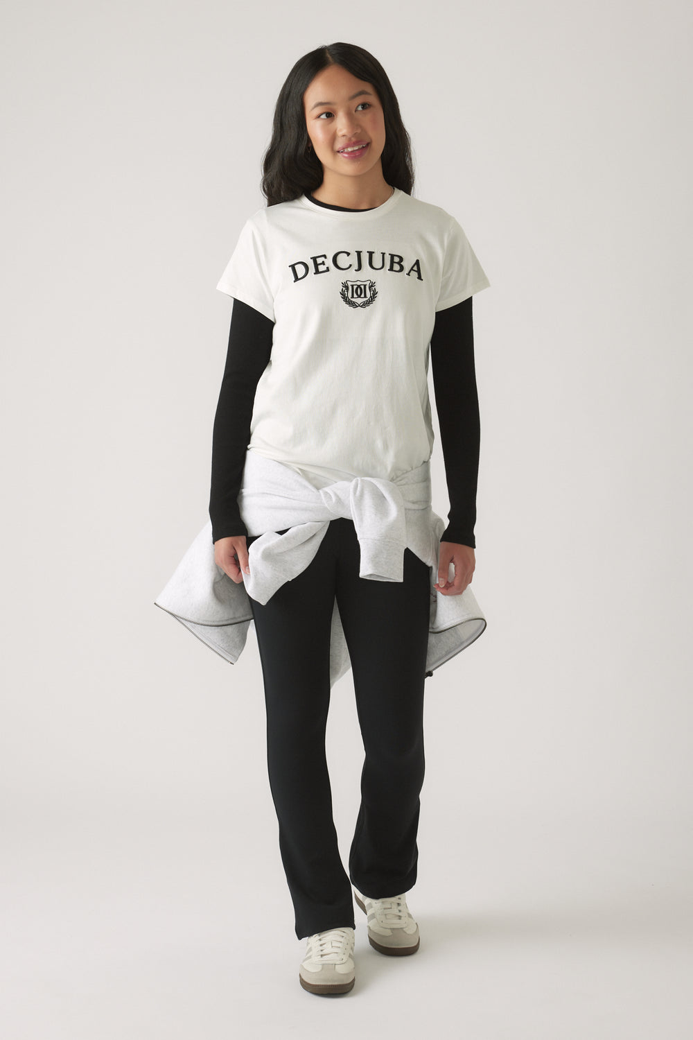 Girl wearing a white logo tee layered over a black long-sleeve top, paired with black pants and sneakers, with a tied sweatshirt around the waist.