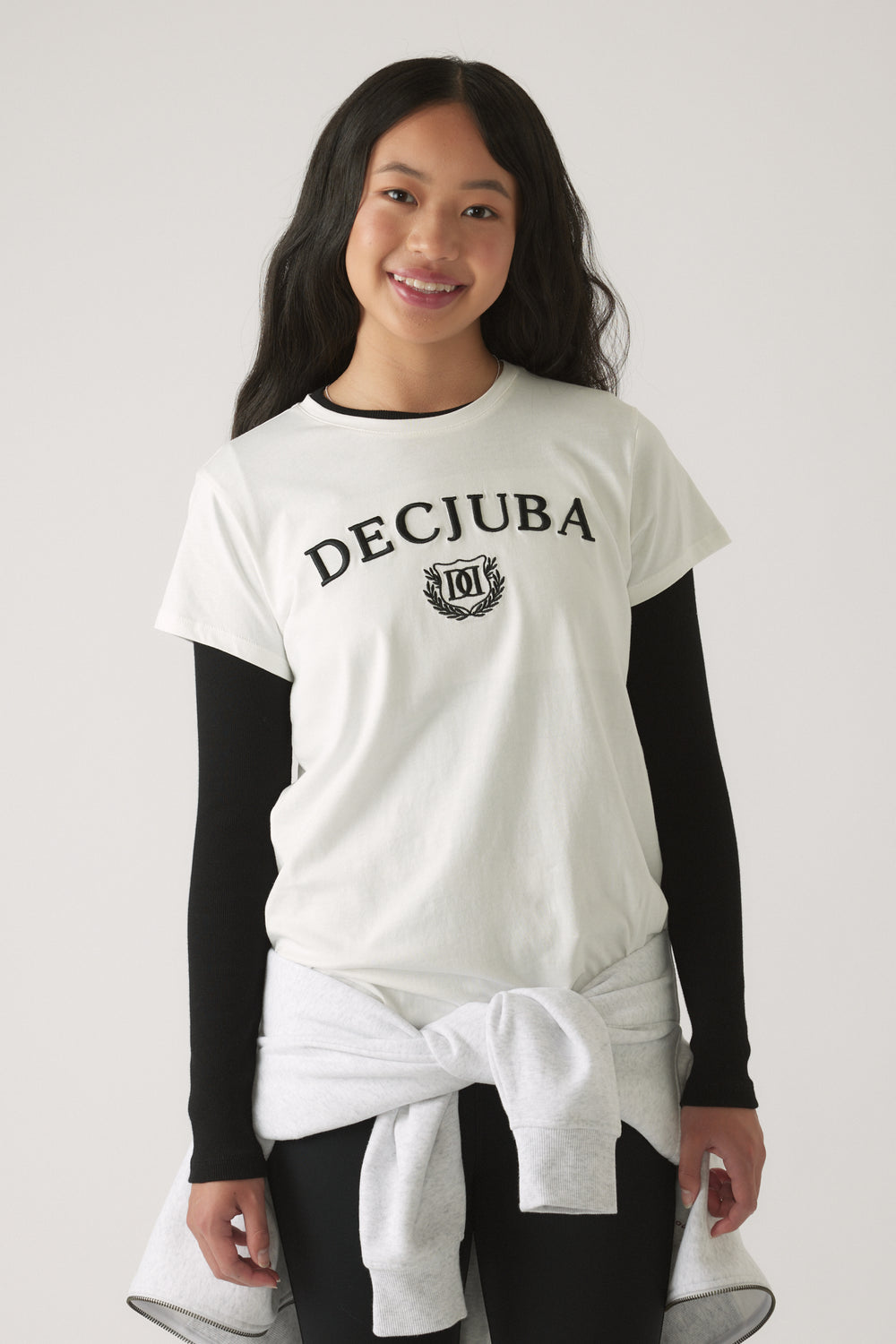 Teen girl wearing a white graphic tee layered over a black long-sleeve top, paired with a grey sweatshirt tied at the waist.