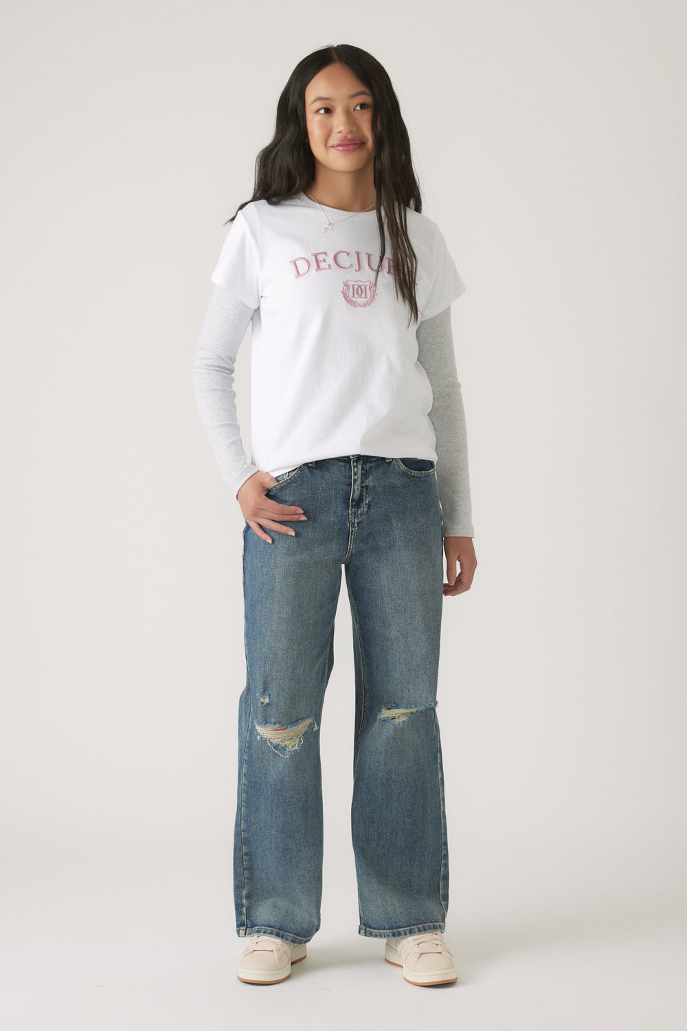 Girl wearing a white Girls Decjuba Heritage Tee layered over a long sleeve grey top with relaxed distressed jeans