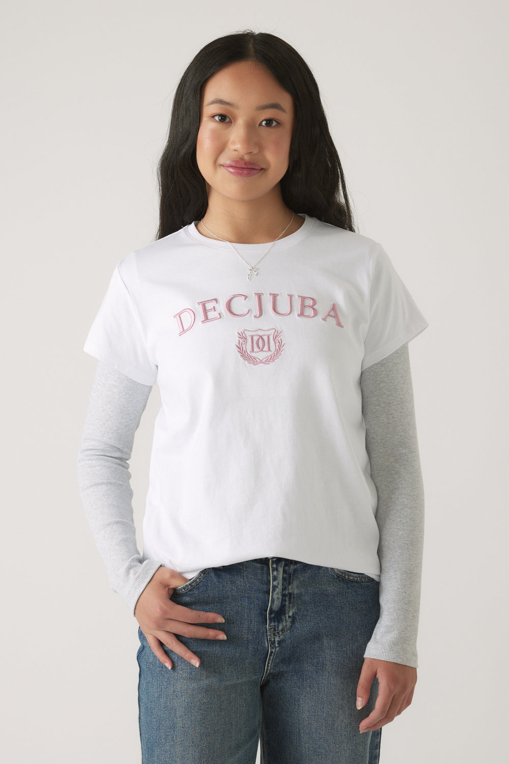Girl wearing a white Girls Decjuba Heritage Tee with pink lettering layered over grey long sleeves and blue denim jeans