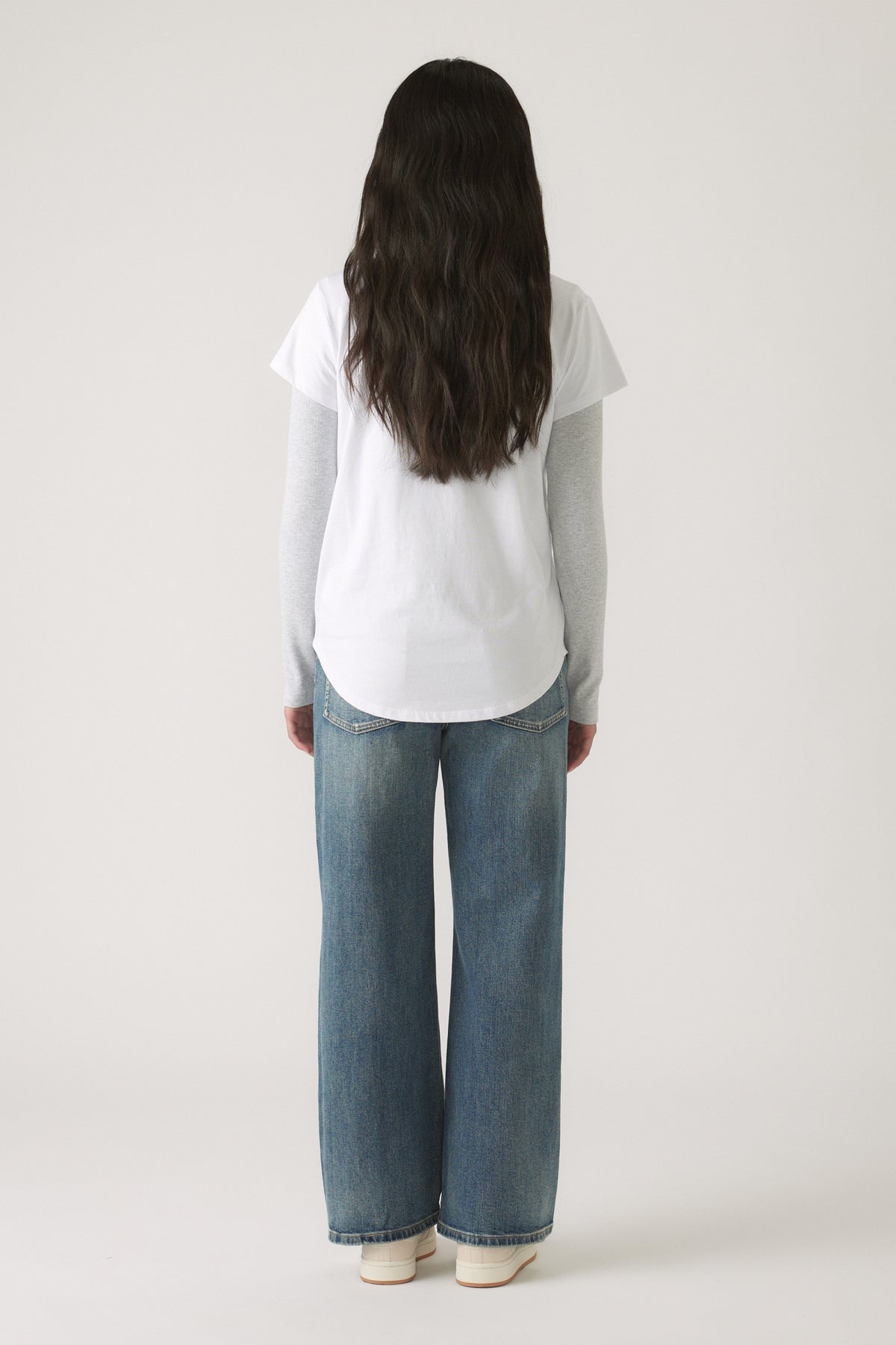 Girl seen from behind wearing a Girls Decjuba Heritage Tee layered over a long sleeve top with relaxed wide-leg jeans and sneakers